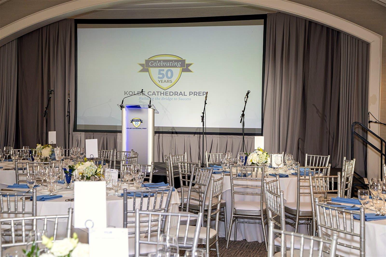 The ballroom set for dinner before guests arrived: round tables, silver chairs, and the “Celebrating 50 Years” crest on the screen behind the podium.