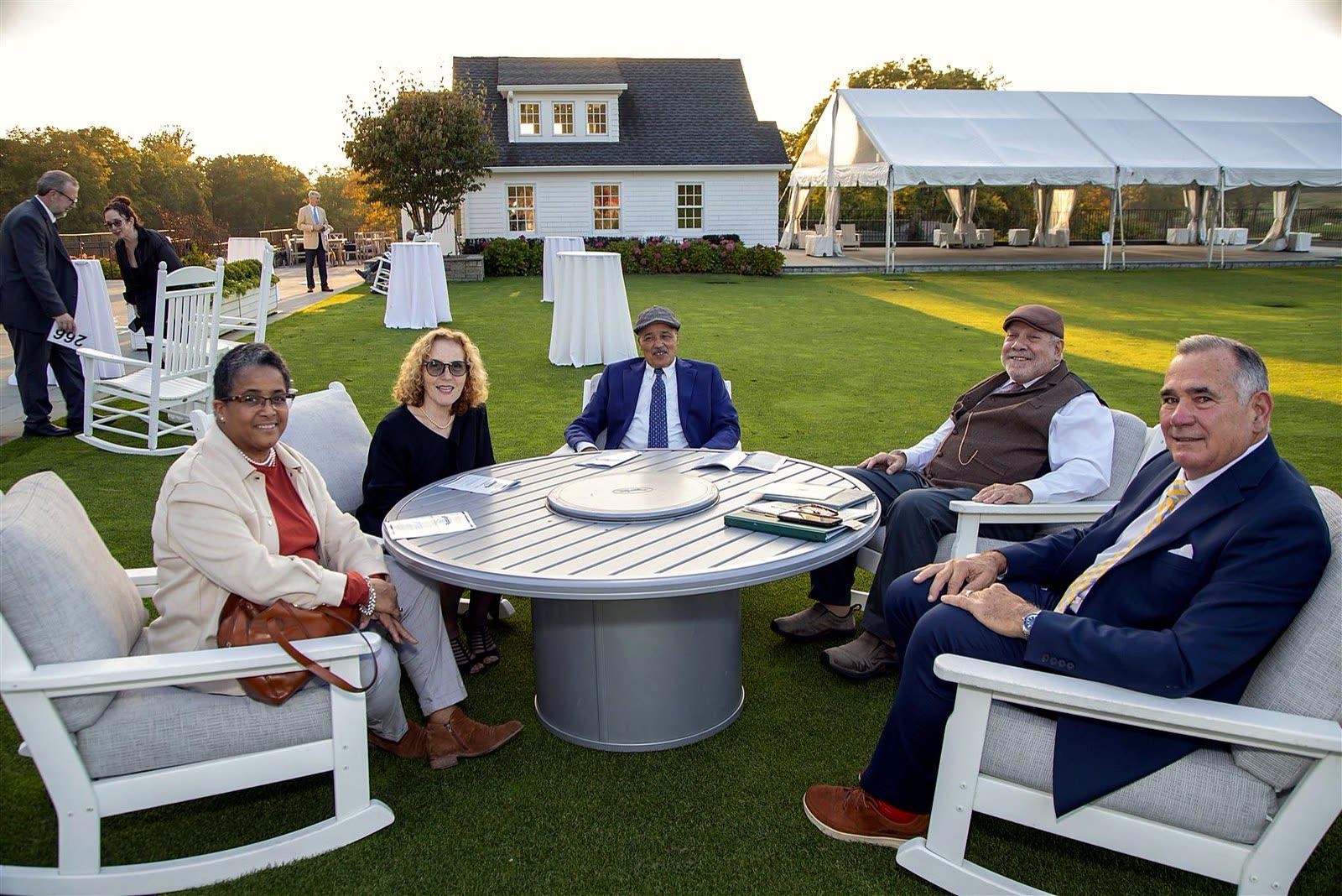 Five guests relaxing in white rocking chairs around a round table on the lawn at sunset, with the clubhouse and a white tent behind them.