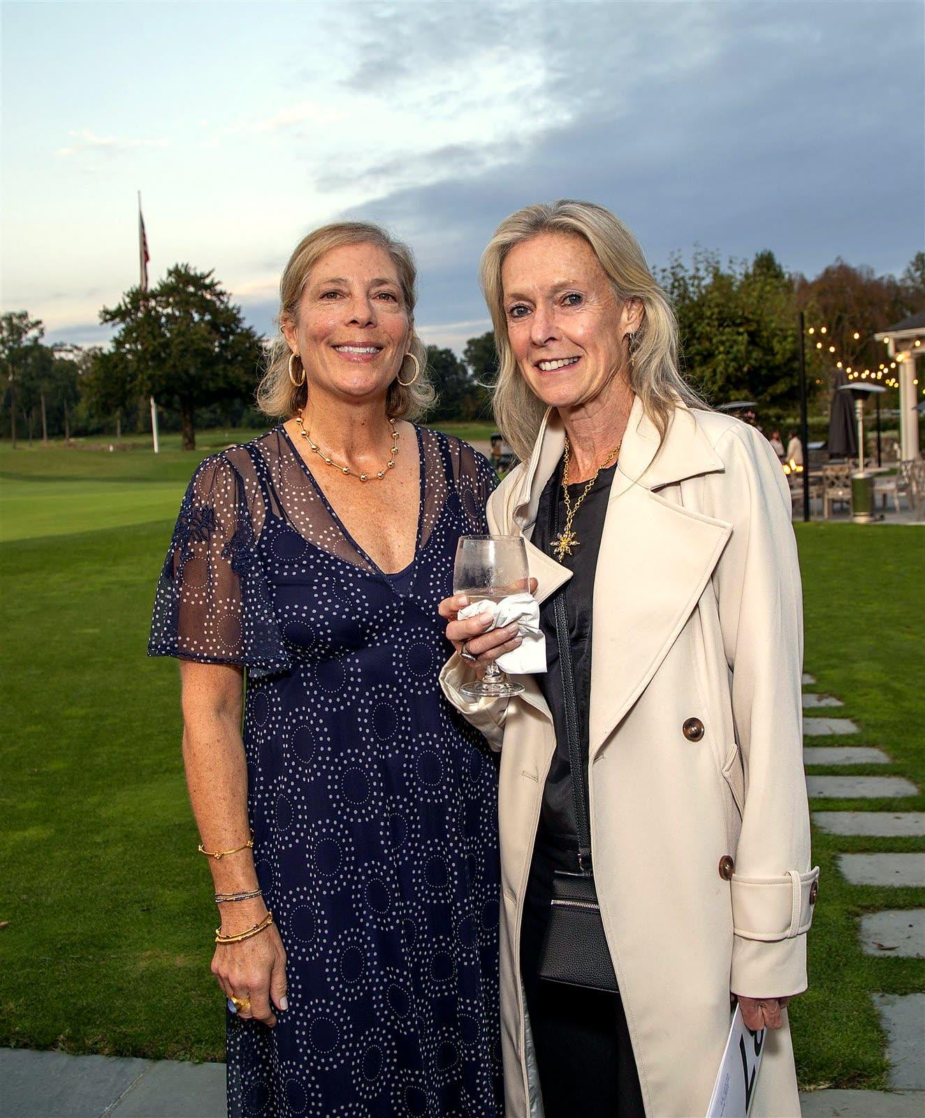Two guests smiling on the lawn at dusk, one holding a glass.