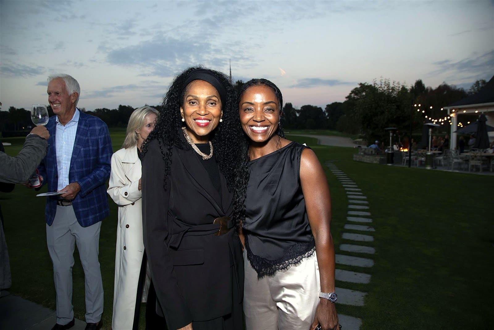 Two guests posing shoulder to shoulder on the lawn at dusk as others mingle under string lights behind them.