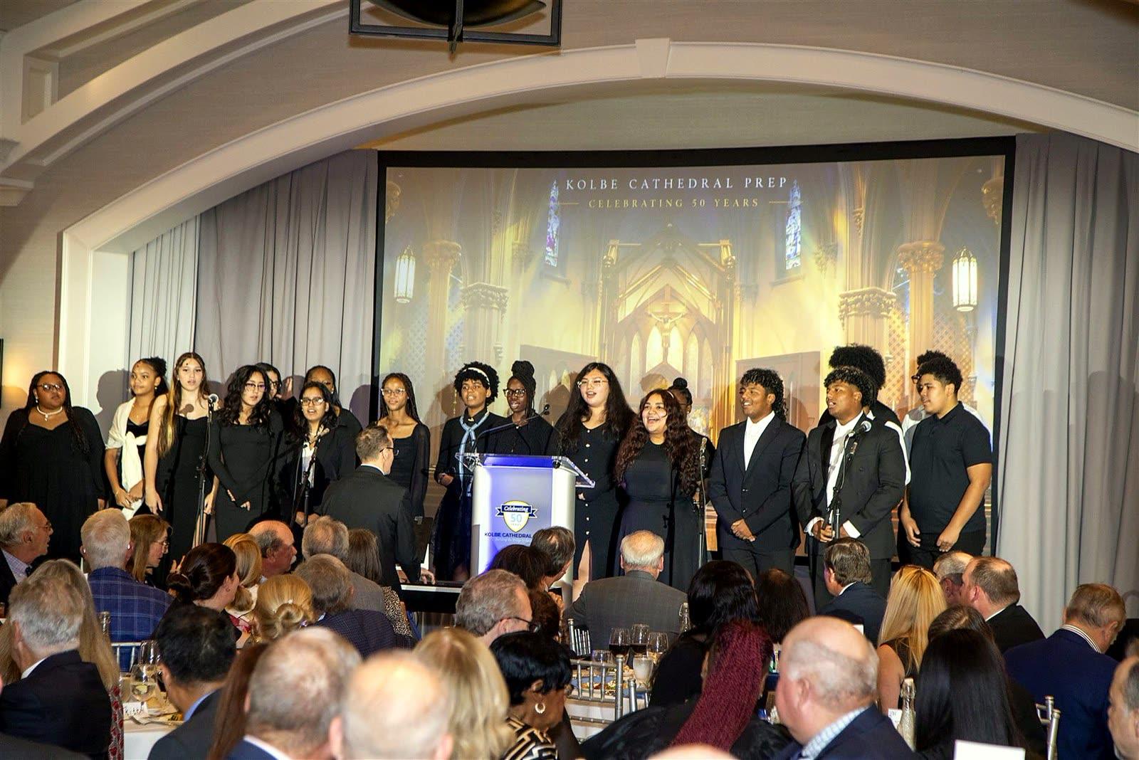 The Kolbe student choir on stage in front of a stained-glass cathedral projection, facing a full ballroom.