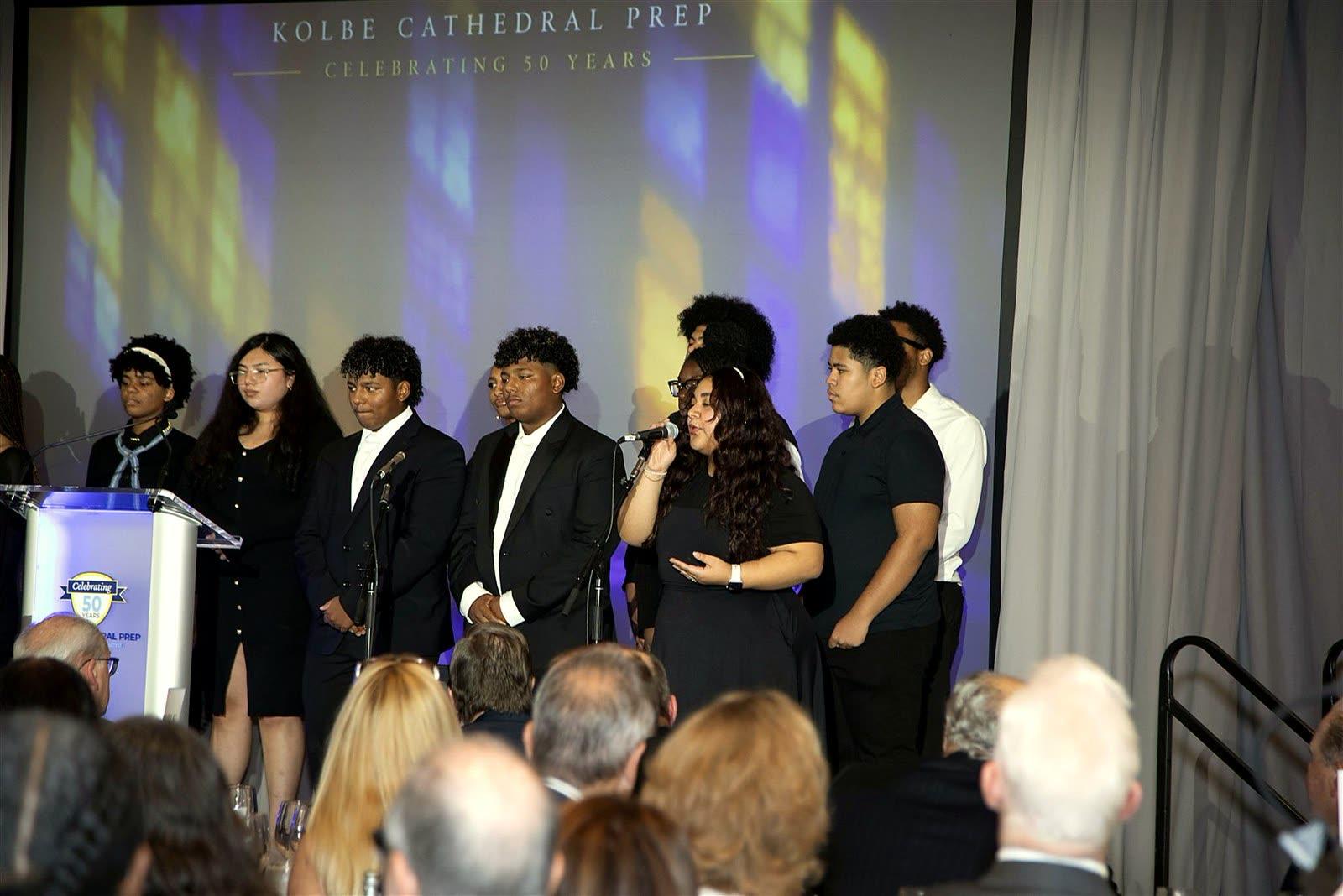 Kolbe students singing at microphones on stage as guests watch.