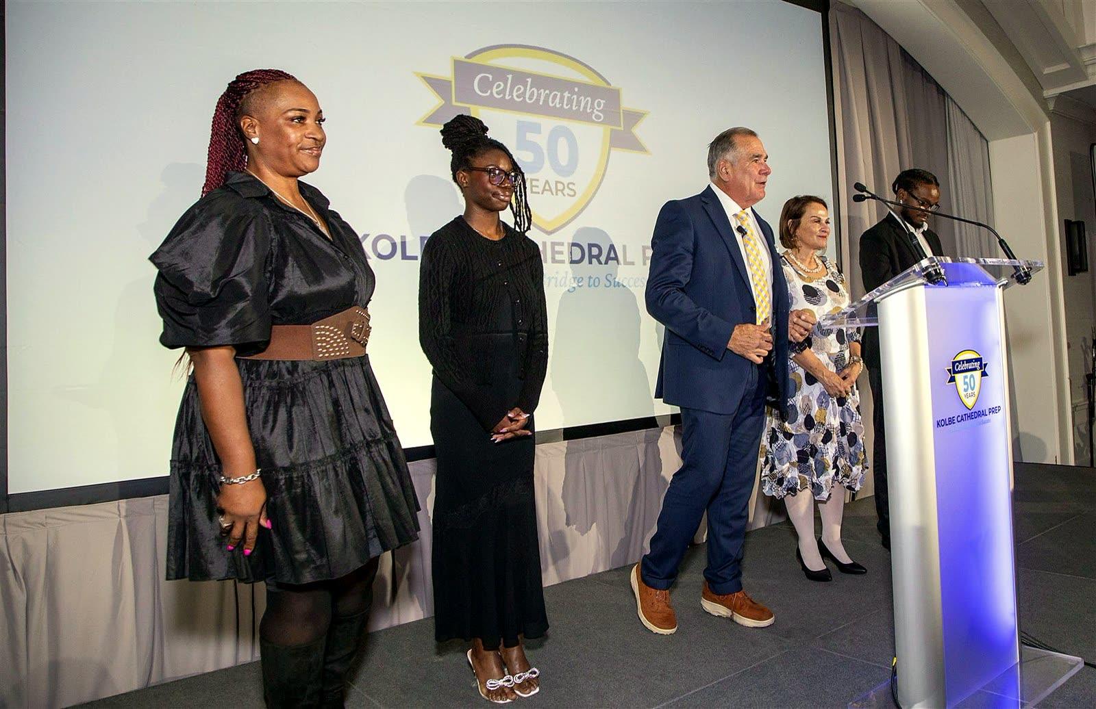 Five people on stage, one speaking beside the podium, in front of the “Celebrating 50 Years” crest.